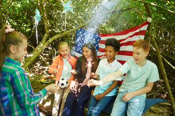 Multi-ethnic group of kids lighting sparklers while hiding under branches of big tree in forest or...