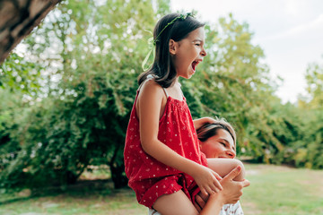 Smiling mother playing and riding on her shoulder her daughter in the park. Happy kid having fun with her mom enjoying the time together. Mother and child share love with each other.