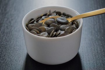 Raw pumpkin seeds and spoon in white bowl on black table.