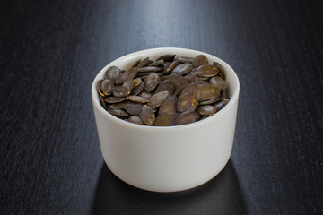 Raw pumpkin seeds and spoon in white bowl on black table.