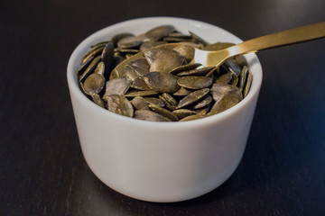 Raw pumpkin seeds and spoon in white bowl on black table.