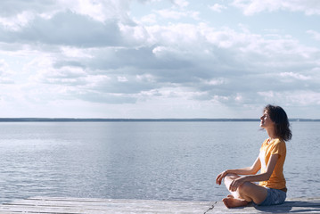 Side view of A young pretty woman enjoys the rest, sitting on the shore of the river in lotus position