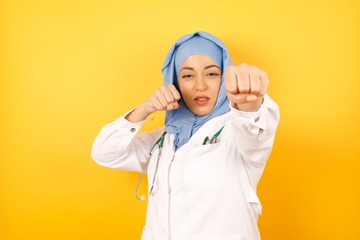 young beautiful Arab doctor woman wearing medical uniform standing over yellow background Punching fist to fight, aggressive and angry attack, threat and violence