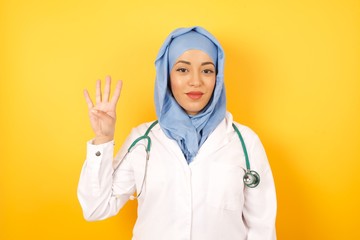 young beautiful Arab doctor woman wearing medical uniform standing over yellow background showing and pointing up with fingers number four while smiling confident and happy.