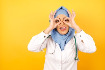 Young arab doctor woman wearing medical uniform standing over yellow background  doing ok gesture like binoculars sticking tongue out, eyes looking through fingers. Crazy expression.