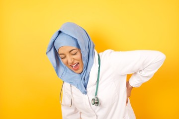 Young arab doctor woman wearing medical uniform standing over yellow background   suffering strong backache, touching back with hand, muscular pain.