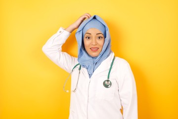 Young arab doctor woman wearing medical uniform standing over yellow background  being confused and...