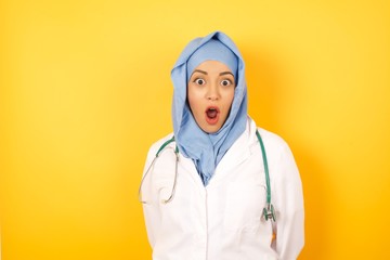 Oh my God. Surprised Young arab doctor woman wearing medical uniform standing over yellow background , stares at camera with shocked expression, exclaims with unexpectedness