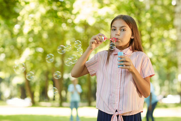 Waist up portrait of teenage girl blowing bubbles and looking at camera while standing in green park outdoors, copy space