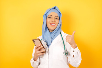 Portrait of  beautiful muslim doctor woman wearing medical uniform  using and texting with smartphone over isolated background happy with big smile doing ok sign, thumb up with fingers, excellent sign