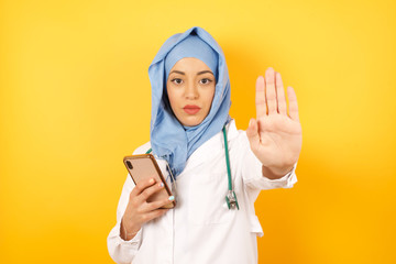 Portrait of beautiful muslim doctor woman wearing medical uniform  using and texting with smartphone over isolated background with open hand doing stop sign with serious and confident expression, 