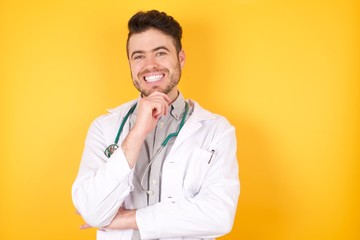 Optimistic Caucasian doctor man, keeps hands partly crossed and hand under chin, looks at camera with pleasure, wears medical uniform, isolated over yellow background. Happy emotions concept.