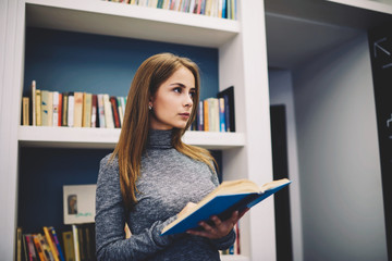 Young attractive female student thinking over plot of detective story while reading book standing...