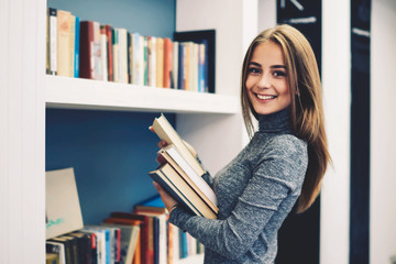 Cheerful female young professional librarian smiling while making revision of books putting them on new bookshelves in alphabetical order working in modern university library for pupils and students