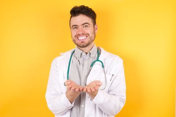 Handsome young Caucasian doctor man wearing medical uniform and stethoscope over isolated yellow background Smiling with hands palms together receiving or giving gesture. Hold and protection