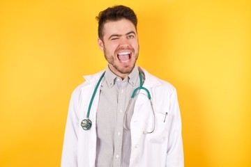 Handsome young Caucasian doctor man wearing medical uniform, over yellow background winking looking at the camera with sexy expression, cheerful and happy face.