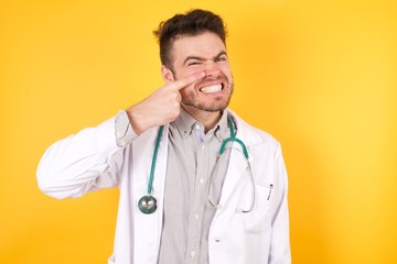 Young Caucasian doctor man wearing medical uniform standing over isolated over yellow background pointing unhappy to pimple on forehead, ugly infection of blackhead. Acne and skin problem