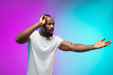 Naklejka premium Shocked asking. African-american young man's portrait on gradient studio background in neon. Beautiful male model in casual style, white shirt. Concept of human emotions, facial expression, sales, ad.