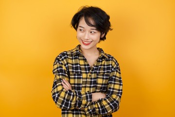 Portrait of dreamy rest relaxed Young asian woman with short hair wearing plaid shirt standing over yellow background crossing arms, looks good 