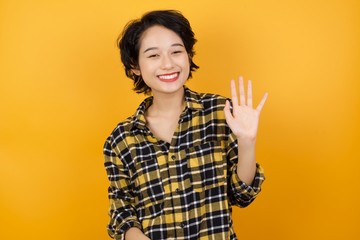 Young asian woman with short hair wearing plaid shirt standing over yellow background Waiving saying hello happy and smiling, friendly welcome gesture.