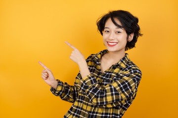 Photo of Young asian woman with short hair wearing plaid shirt standing over yellow background indicating with forefinger empty space showing best low prices 