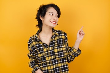 Smiling Young asian woman with short hair wearing plaid shirt standing over yellow background pointing up and looking at the camera 
