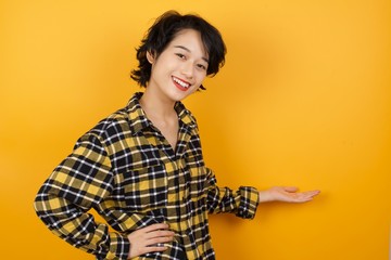 Young asian woman with short hair wearing plaid shirt standing over yellow background feeling happy and cheerful, smiling and welcoming you, inviting you in with a friendly gesture 