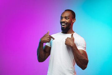 Pointing, showing. African-american young man's portrait on gradient studio background in neon. Beautiful male model in casual style, white shirt. Concept of human emotions, facial expression, sales