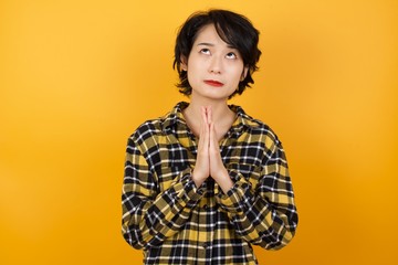 Young beautiful Asian woman wearing plaid shirt over yellow background begging and praying with hands together with hope expression on face very emotional and worried. Asking