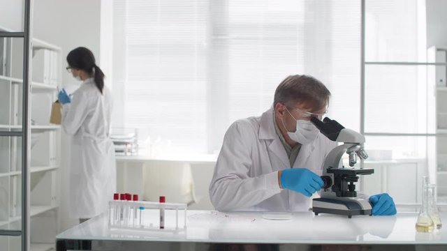Medium shot of male biomedical scientist in gloves, goggles and face mask sitting at desk in laboratory and looking into microscope. Female scientist in lab coat looking for document in background