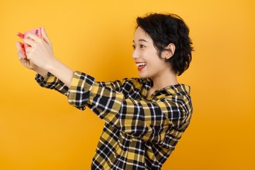 Close-up portrait Young asian woman with short hair wearing plaid shirt standing over yellow background taking a selfie to post it on social media or having a video call with friends.