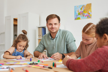 Portrait of male teacher smiling at camera while working with multi-ethnic group of children...