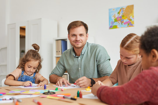 Portrait of male teacher smiling at camera while working with multi-ethnic group of children drawing pictures during art class in school or development center, copy space