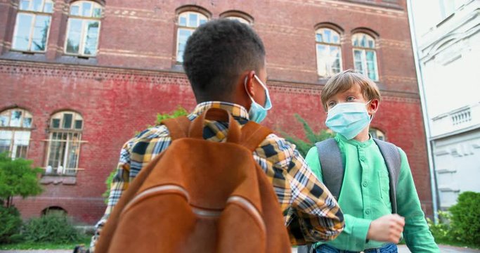 Close up portrait of multi-ethnic children in medical masks greeting with elbows near school. Cute African American and Caucasian boys standing outdoors with schoolbags. Pupils life Quarantine concept