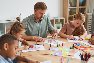 Portrait of male teacher working with multi-ethnic group of children drawing pictures during art...