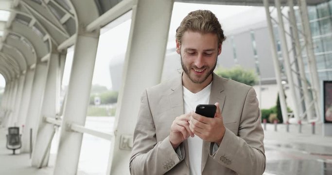 Caucasian Businessman is standing in Industrial Quartal and browsing his Phone. Handsome Young Businessman is having Good Mood, looking satisfied. Businessman is walking in Raing, smiles. People.