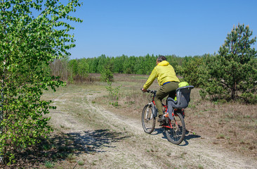 Fototapeta na wymiar Dad in bright clothes rides his little daughter in a bicycle seat on a bicycle along a dirt road through the woods