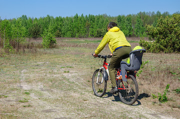 Dad in bright clothes rides his little daughter in a bicycle seat on a bicycle along a dirt road through the woods