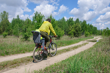 Naklejka premium Dad in bright clothes rides his little daughter in a bicycle seat on a bicycle along a dirt road through the woods