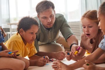 Portrait of young male teacher working with kids drawing pictures while having fun in pre school or...