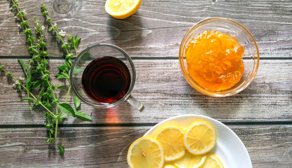 A cup of tea, a sprig of mint, orange jam, lemon on a plate on a dark wooden background. Top view