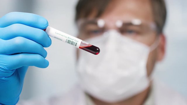 Close up with rack focus of male scientist in protective goggles and face mask looking at blood sample in test tube