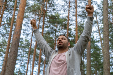 Young handsome man wearing casual clothes, pink shirt and gray sweat shirt celebrating victory with happy smile and winner expression with raised hands in pine tree woods.