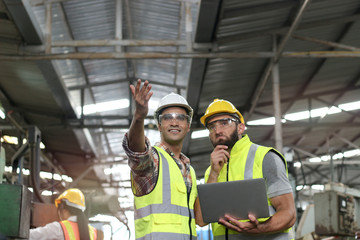 Industrial engineer wearing helmet and safe glasses, holding laptop computer for operating machinery at manufacturing plant factory, worker team working together in industry concept