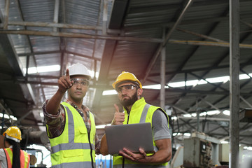 Industrial engineer wearing helmet and safe glasses, holding laptop computer for operating machinery at manufacturing plant factory, worker team working together in industry concept