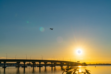 Sweeping lines of Tauranga Harbour Bridge over calm blue water with glow of rising sun at far end as passenger plane take-off and passes over.