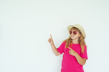 Obraz na płótnie Canvas girl in a bright pink T-shirt, straw hat and sunglasses against a white wall points to the side. copy space..