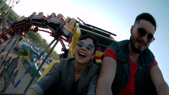 Selfie video of couple riding the roller coaster. Hipster man and stylish woman have fun at amusement park rides. Friends extreme riding, laughing and screaming of excitement, adrenalin.