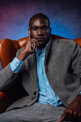A serious well-dressed man seats on chair. Multicolored background. African stylish man with glasses looking at camera with foggy background behind.