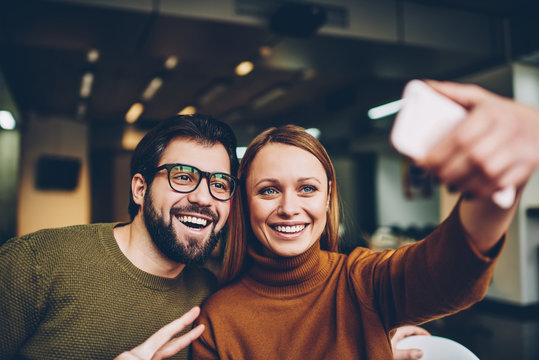Technology and communication, handsome bearded man having fun showing fingers 'peace' on photo while attractive smiling woman clicking selfie using application on modern mobile phone indoors